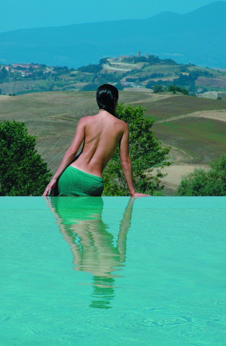 Topless woman sitting at the edge of an infinity swimming pool, looking over Tuscany